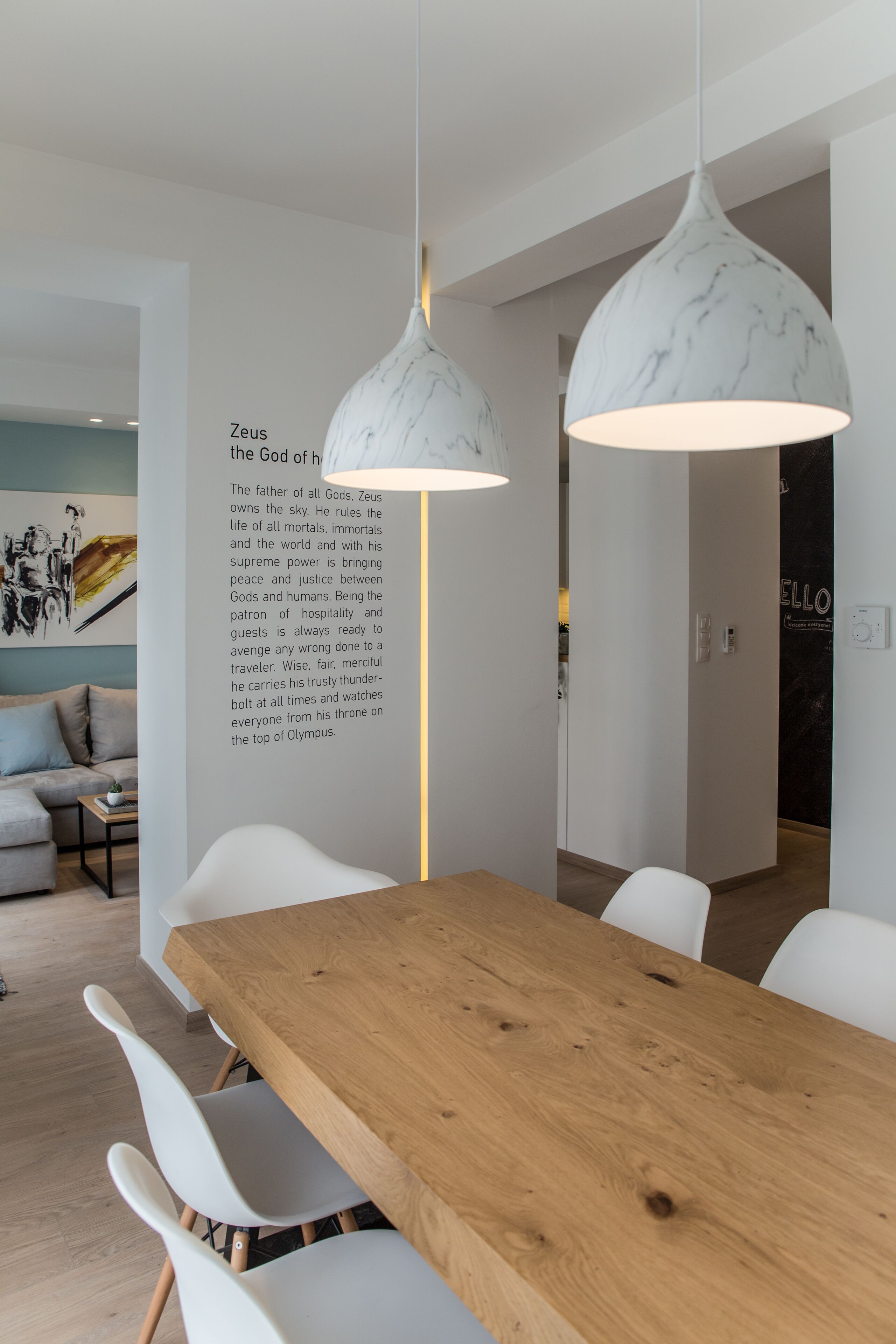 A modern dining area with a wooden table and white chairs, a marble pendant light, and a wall with a text description of Zeus.