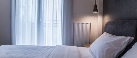Una cama bien hecha con sábanas blancas, una mesita de noche con una lámpara y una ventana con cortinas traslúcidas.