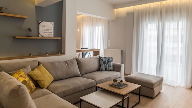 Una sala moderna con sofá, reposapiés y mesa de centro, estante montado en la pared con objetos decorativos y una ventana con cortinas translúcidas.