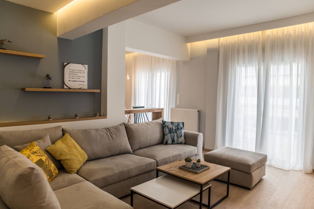 A modern living room with a sofa, ottoman, and coffee table, a wall-mounted shelf with decorative items, and a window with sheer curtains.