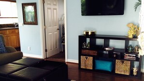 Una sala de estar con un sofá de cuero negro, un televisor de pantalla plana montado en la pared, una mesa de centro de madera y una alfombra estampada.
