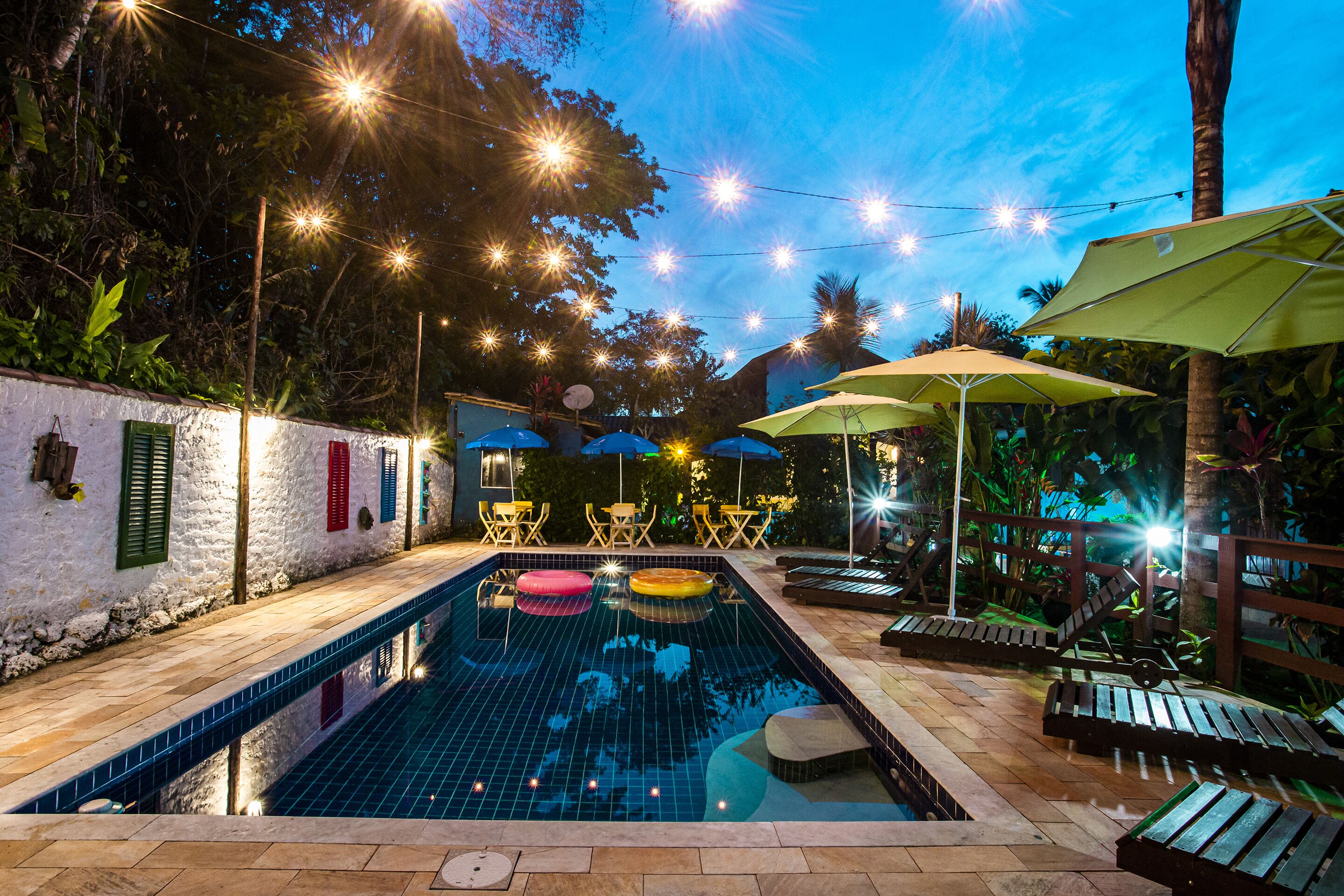A swimming pool with a tiled deck, surrounded by lounge chairs and umbrellas, and string lights overhead.