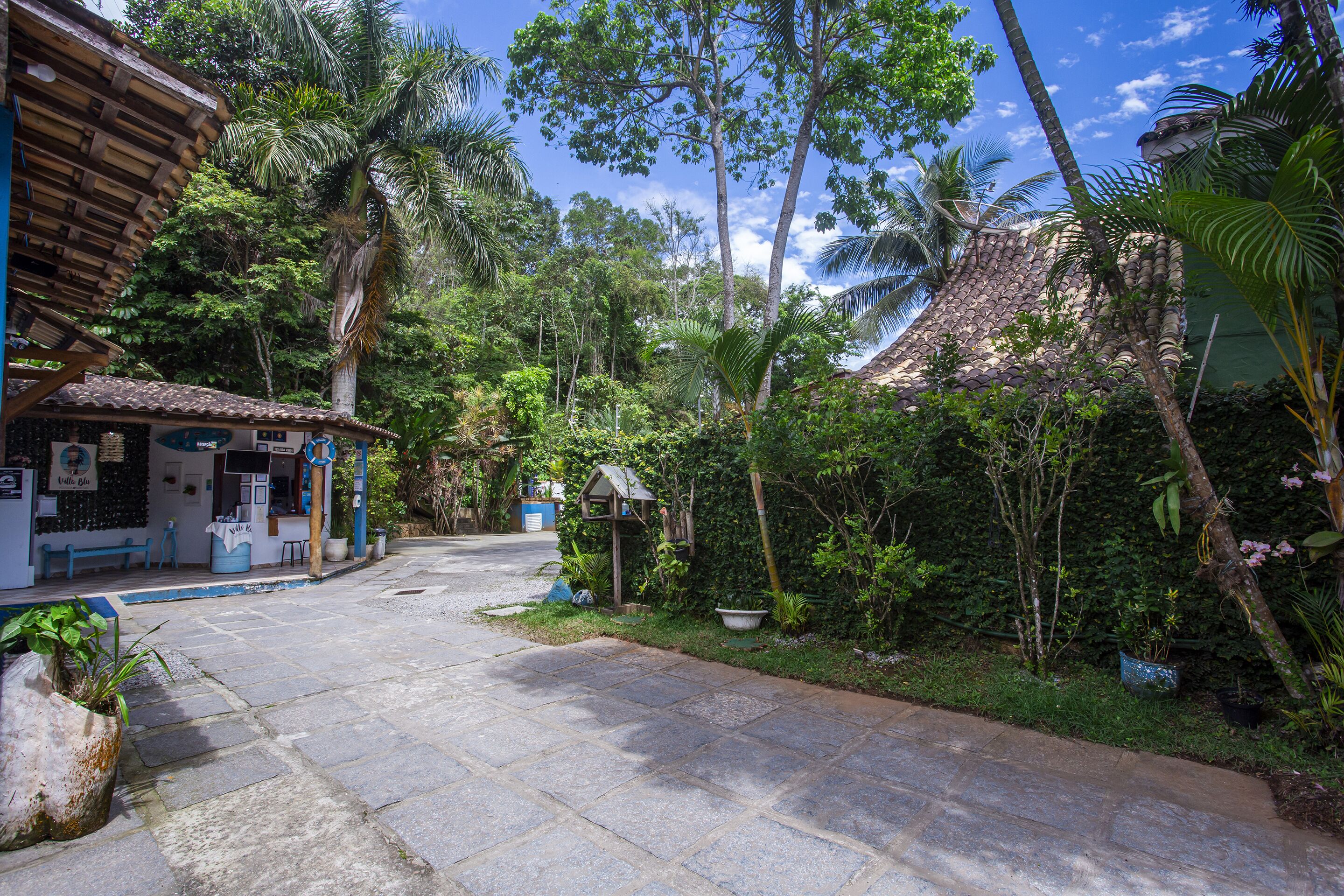 A paved pathway leading to a building with a tiled roof, surrounded by lush greenery and palm trees.