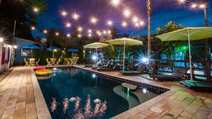 Une piscine entourée de palmiers et illuminée de guirlandes lumineuses, avec des chaises longues et des parasols à proximité.