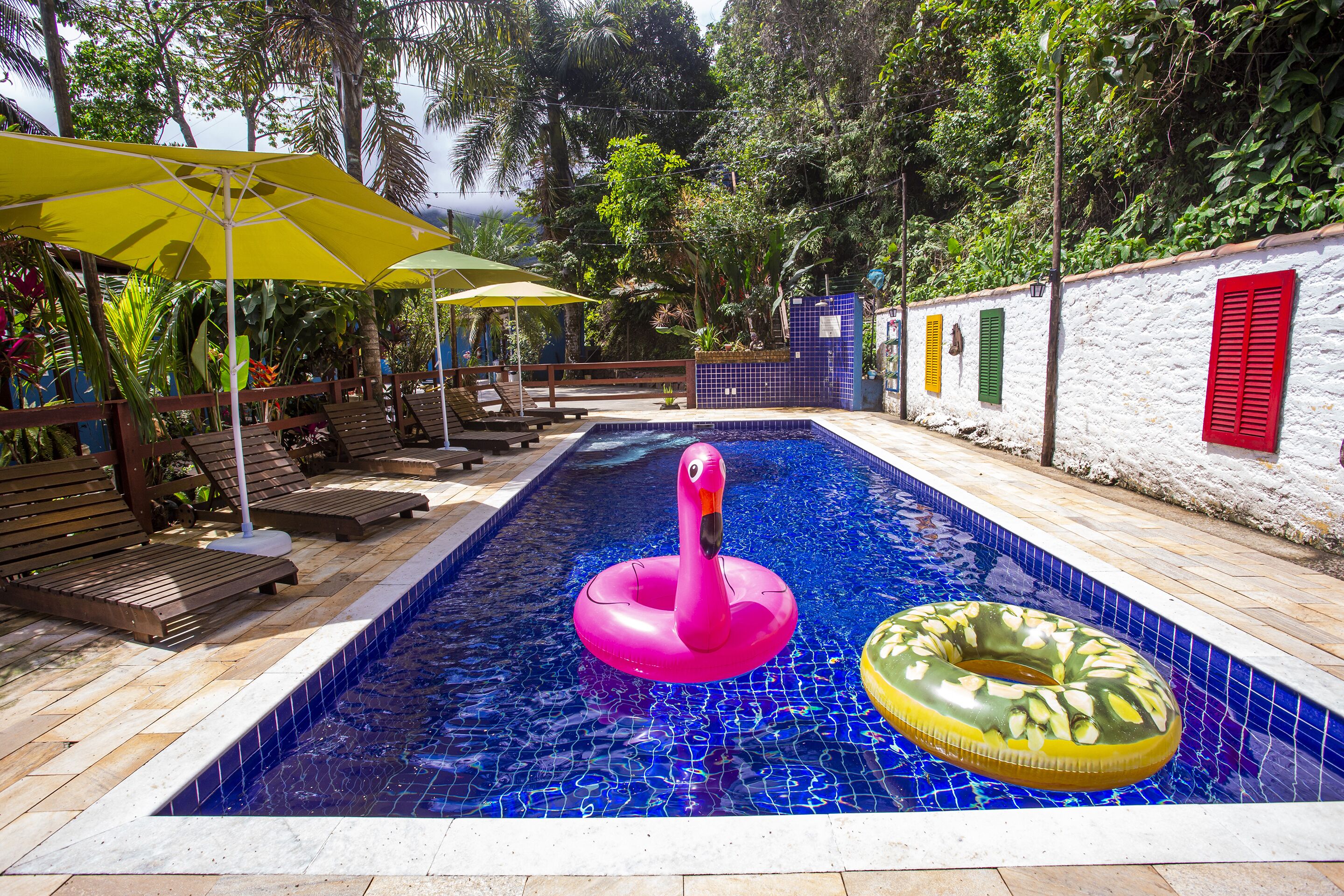 A swimming pool with a pink flamingo float, a yellow and green inflatable ring, and wooden lounge chairs under yellow umbrellas.