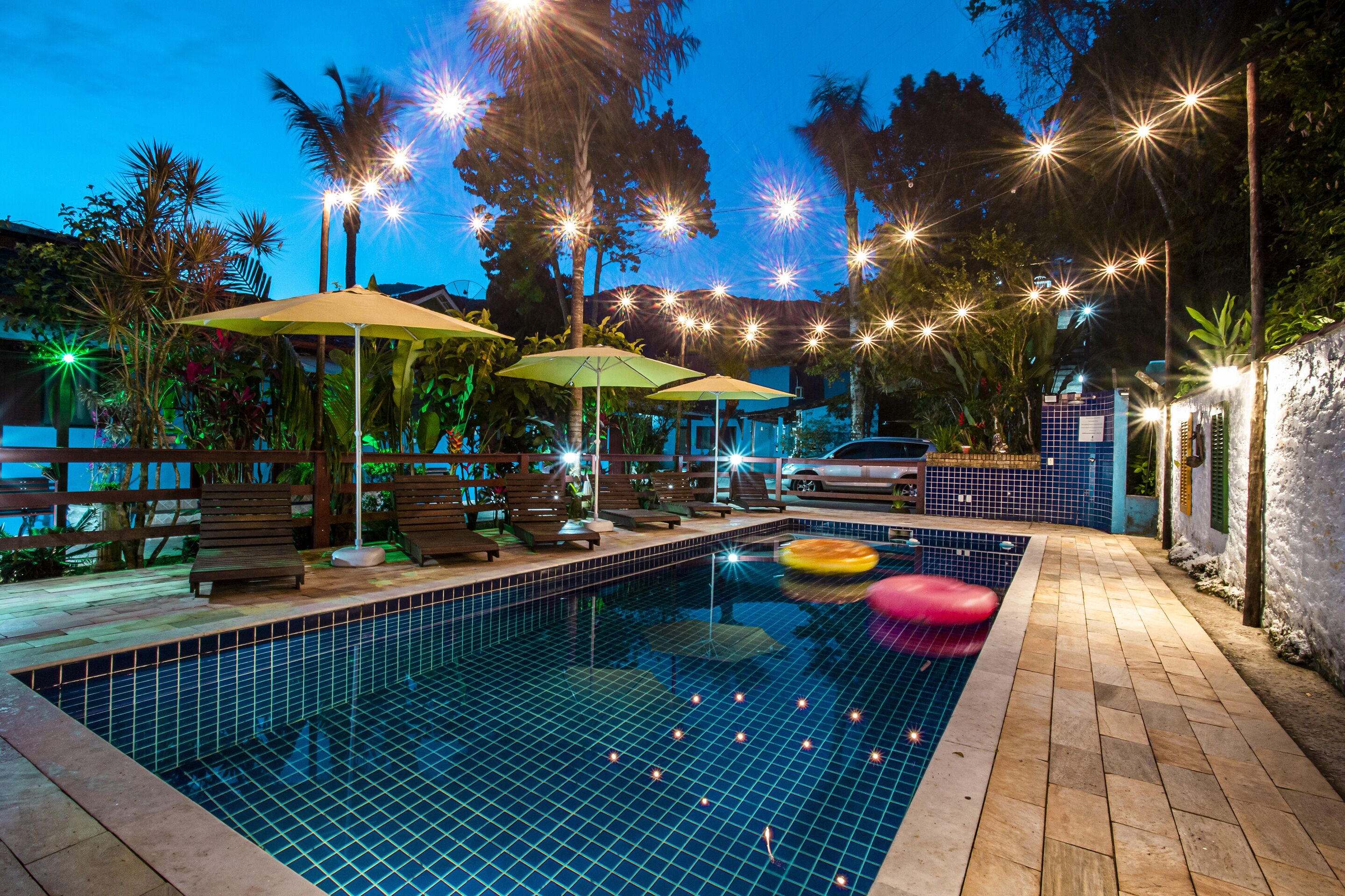 A swimming pool with lounge chairs and umbrellas, surrounded by palm trees and string lights.