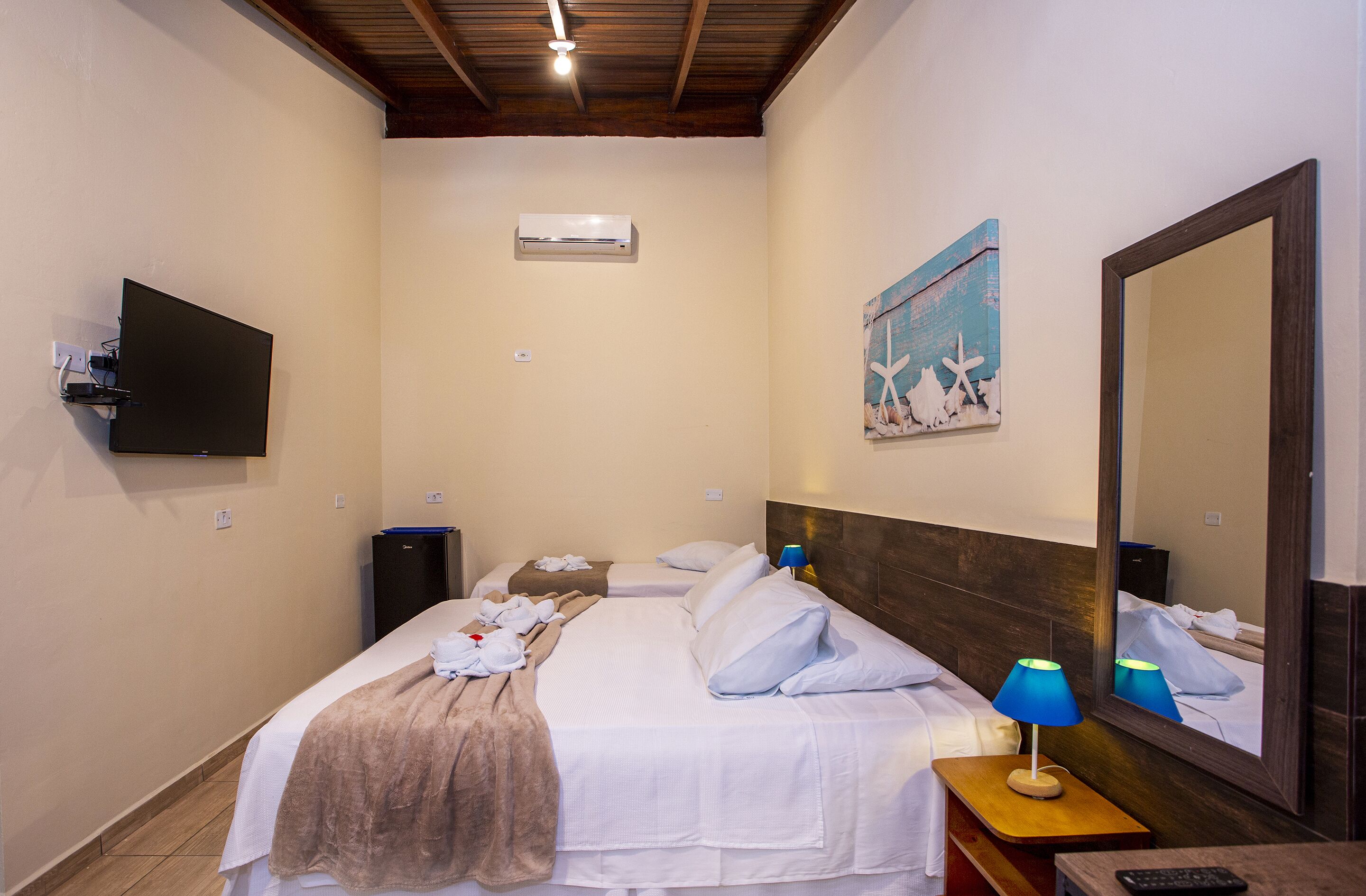 Habitación de hotel con dos camas, un televisor de pantalla plana, aire acondicionado, un cuadro, un espejo y una mesita de noche con lámpara.