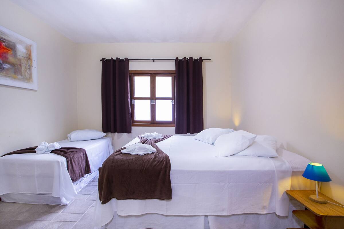 Habitación de hotel con dos camas individuales, una cama grande, una ventana con cortinas, una mesita de noche con lámpara y un cuadro en la pared.