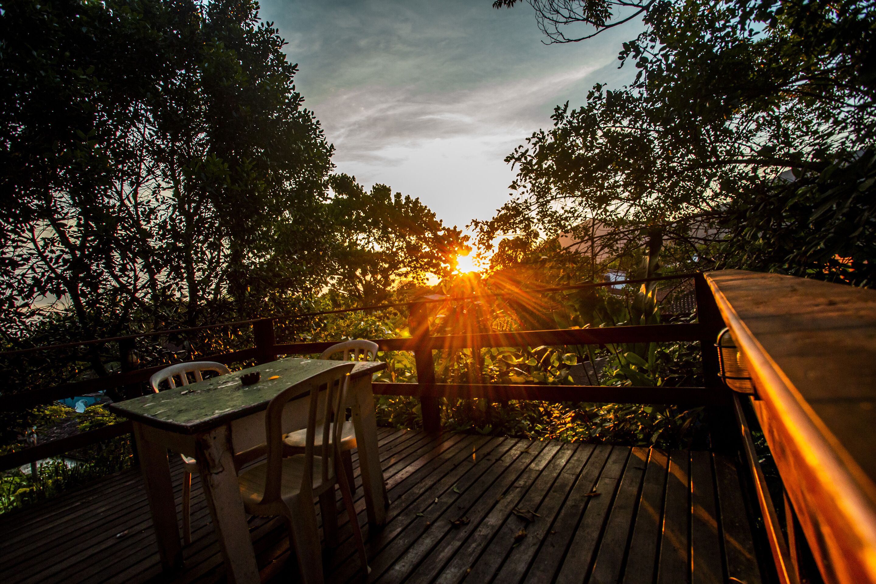 Una terraza de madera con una mesa y sillas, rodeada de árboles, con el sol poniéndose al fondo.