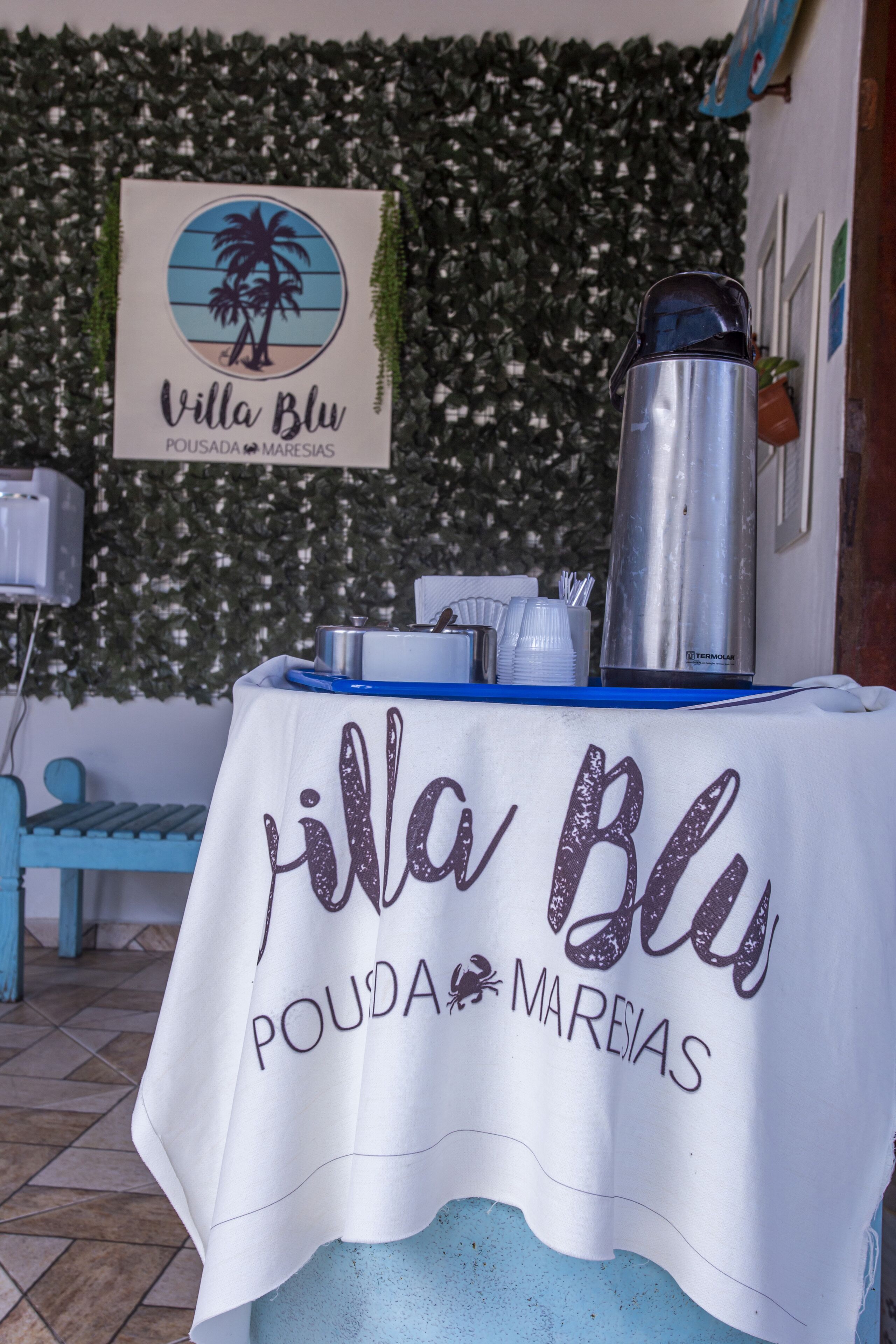 Een tafel met een witte tafelkleed waarop 'Villa Blu POUDA MAREIAS' staat en een roestvrijstalen waterdispenser.