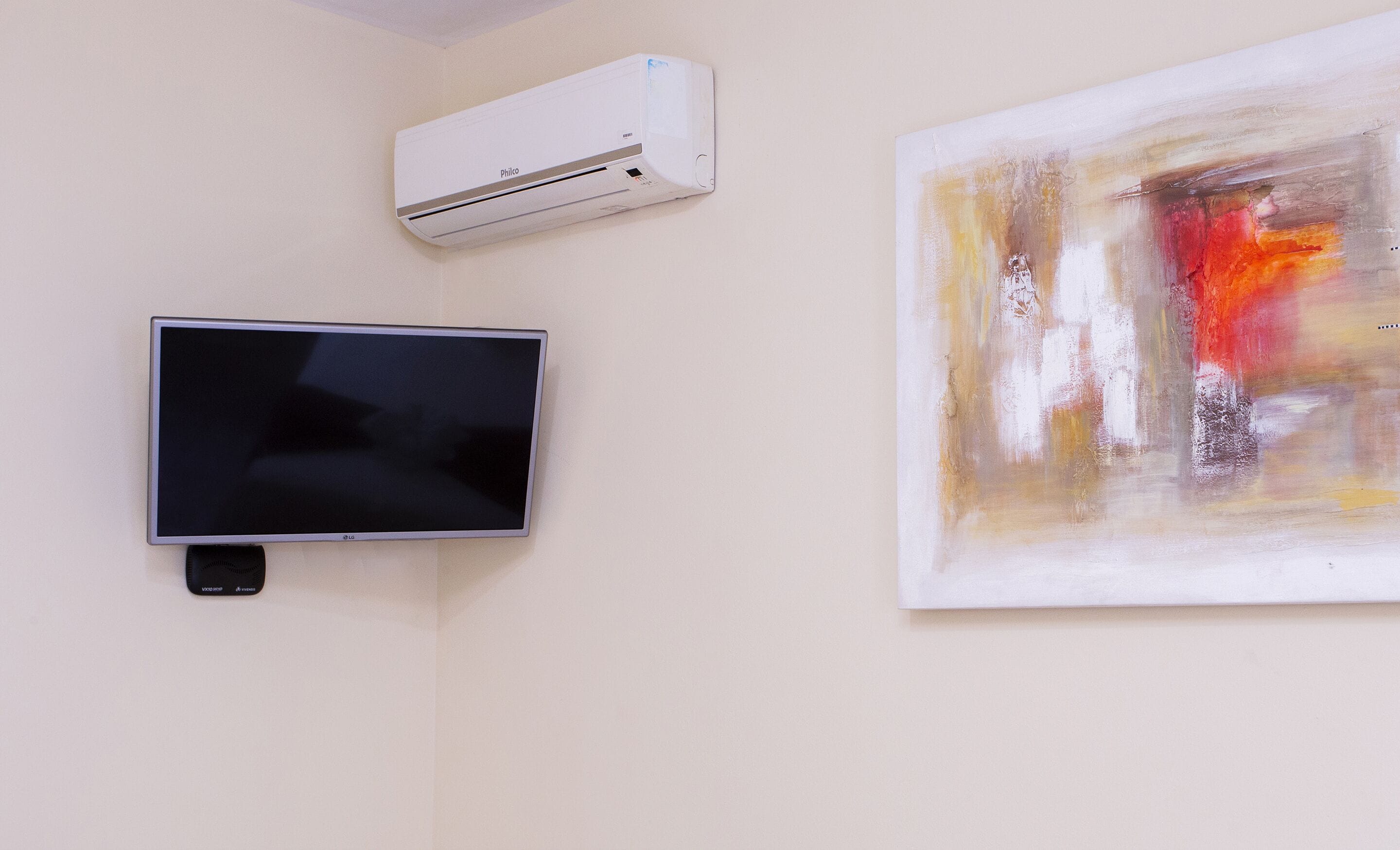 A flat-screen TV mounted on a wall, an air conditioning unit above it, and a large abstract painting to the right.