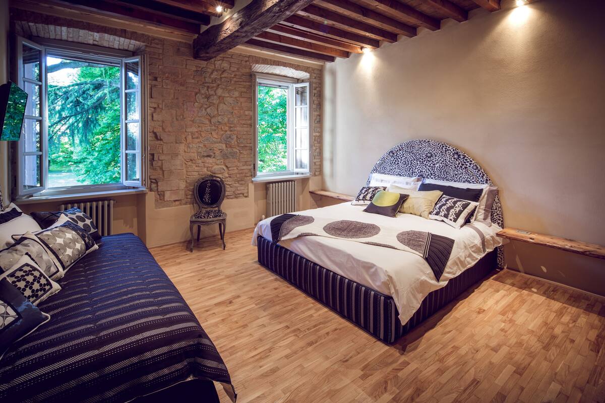 A bedroom with a large bed, a smaller bed, and a chair. There are two windows with views of greenery.