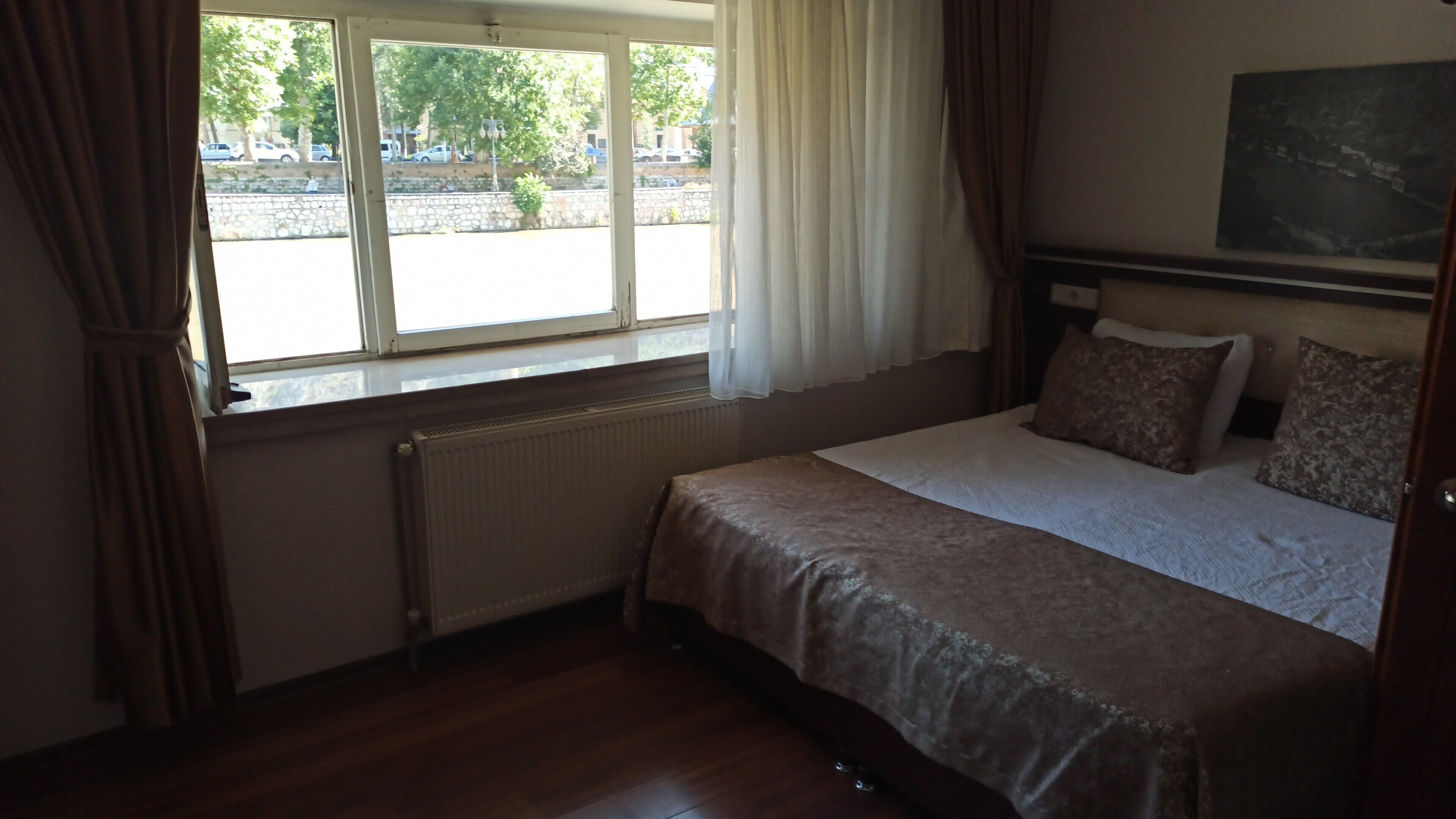 A bedroom with a bed, a radiator, a window with curtains, and a view of the street outside.