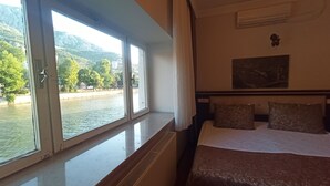 A hotel room with a bed, a window with a view of a river and mountains, and a radiator. - Hasirci Konaklari (Amasya)