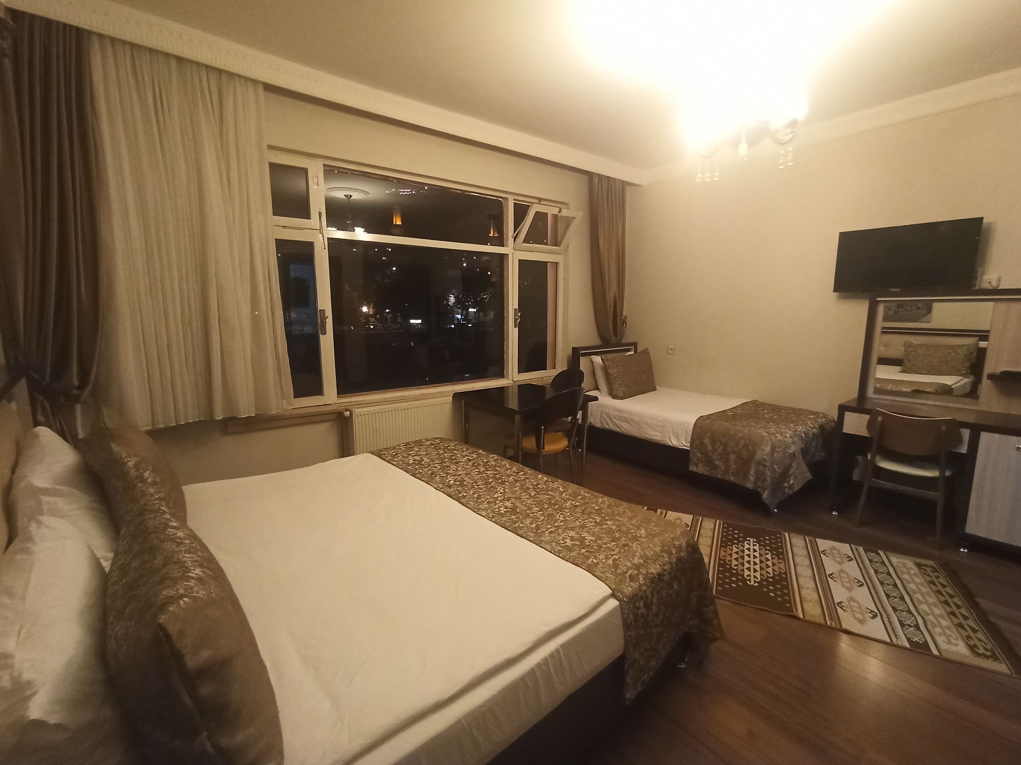 Habitación de hotel con dos camas individuales, un escritorio con silla, televisión y un ventanal con cortinas.