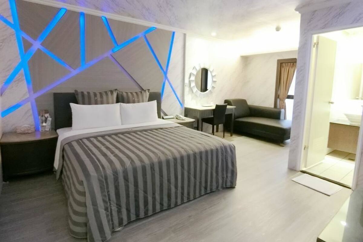 A modern hotel room with a bed, a desk, a sofa, and a bathroom visible through an open door.