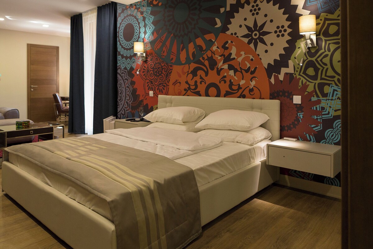 A hotel room with a large bed, a bedside table, a nightstand, and a wall with a colorful abstract design.