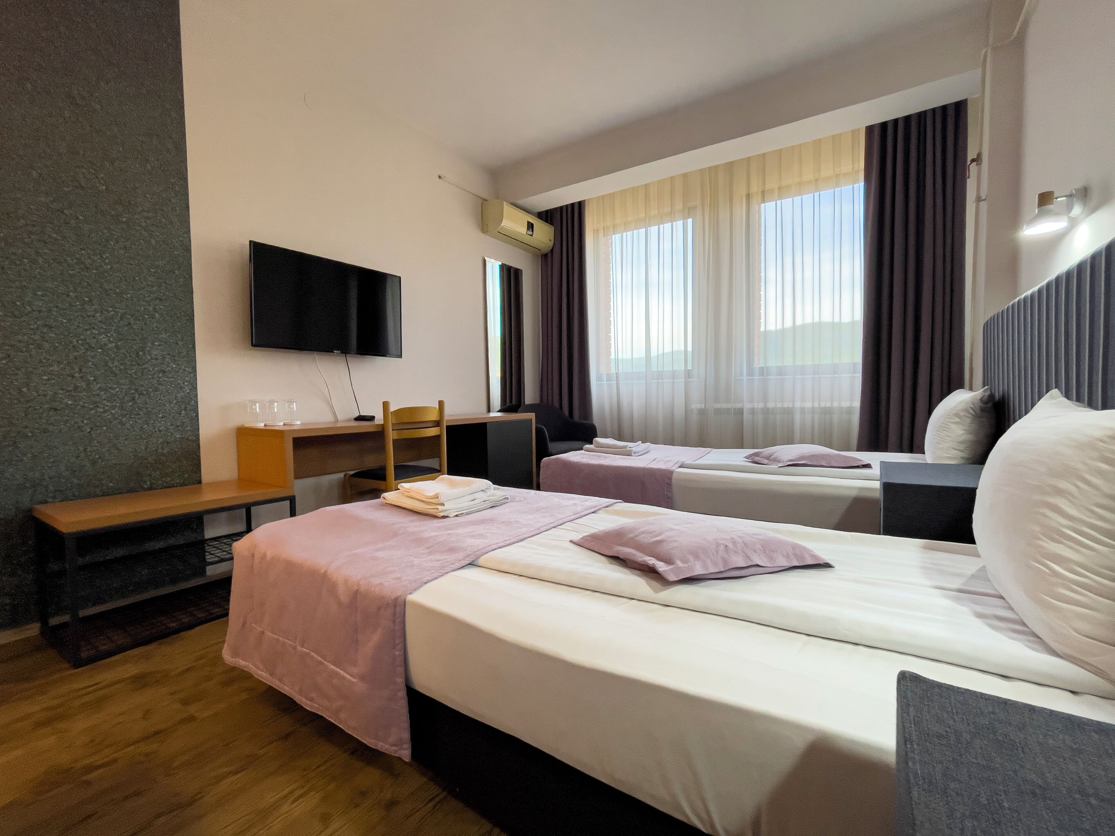A hotel room with two beds, a desk, a chair, a TV mounted on the wall, and a window with curtains.