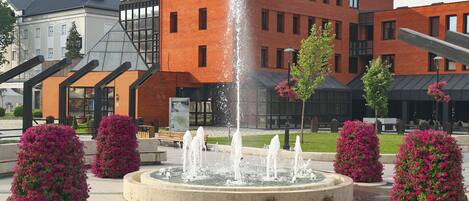 A hotel with a modern brick facade, a central water feature, and surrounding landscaped flower beds.