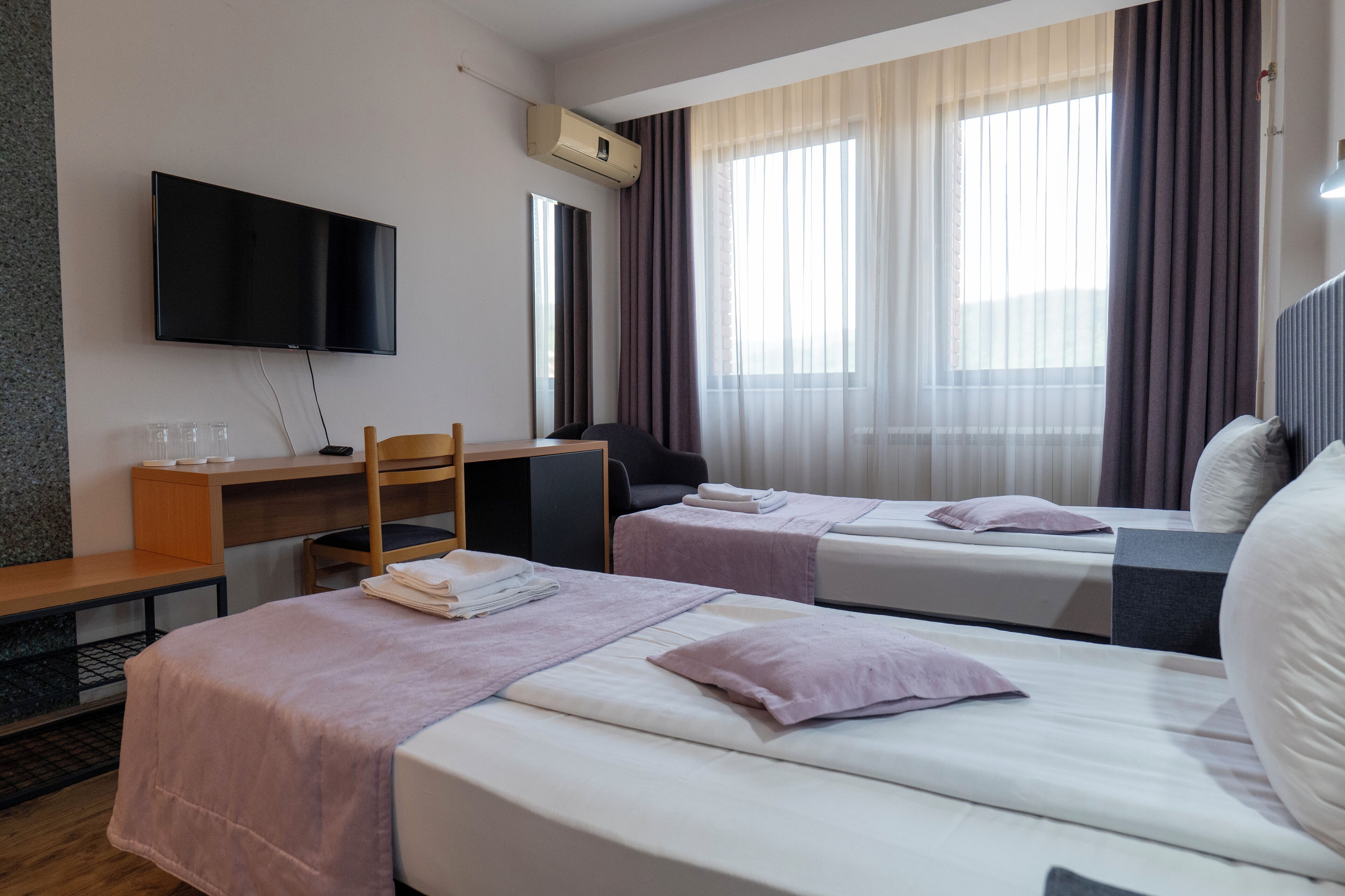 Quarto de hotel com duas camas, uma escrivaninha, uma cadeira, televisão e janela com cortinas.