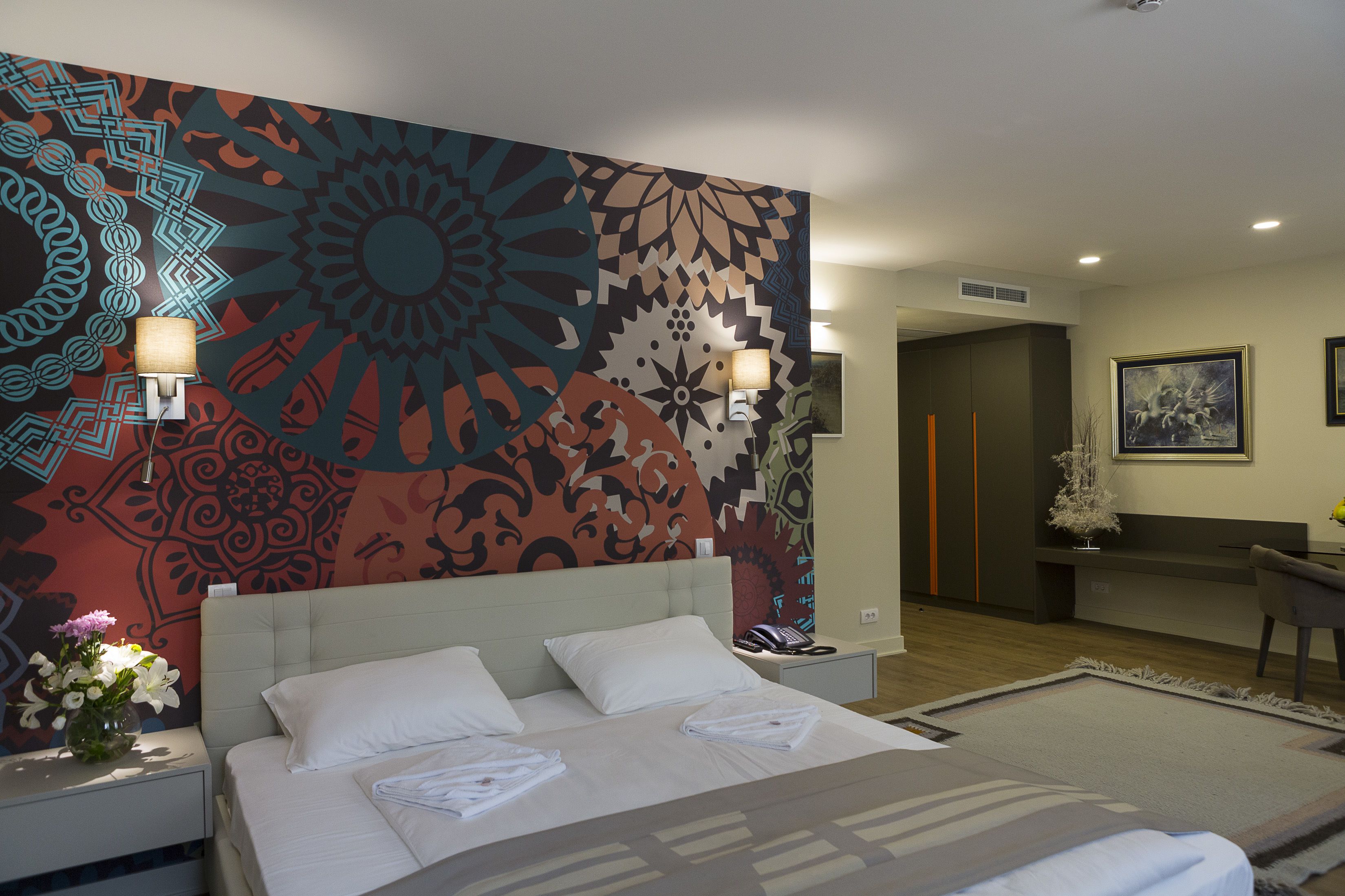 A hotel room with a large wall mural, a bed with white linens, a nightstand with a lamp, and a desk with a chair.