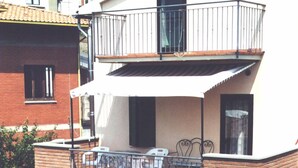 Un edificio de dos pisos con un balcón en el nivel superior. El balcón tiene barandilla metálica y está amueblado con una mesa, sillas y una pequeña planta en maceta.
