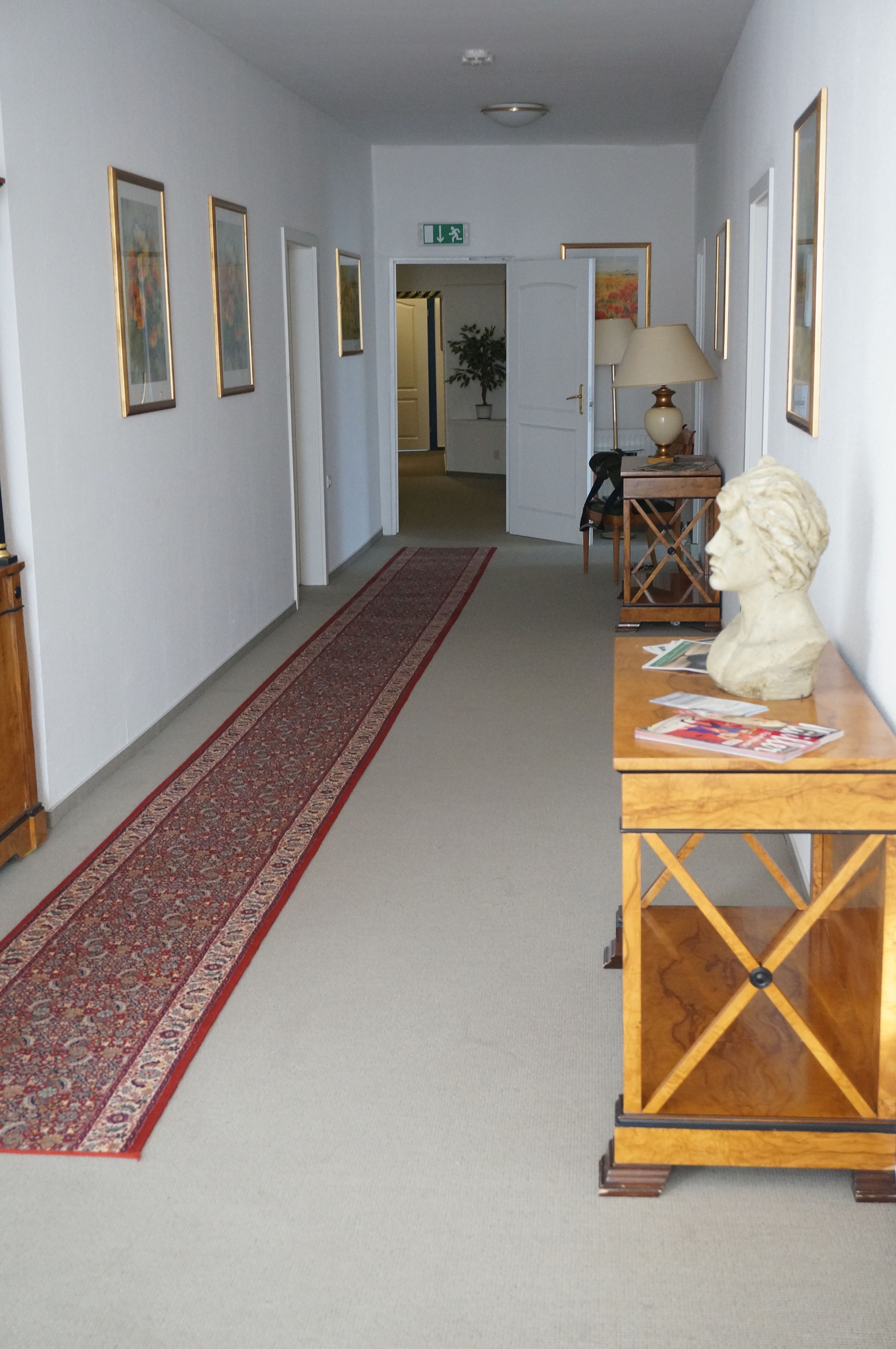 Un pasillo con alfombra roja, cuadros en las paredes, un busto sobre una mesa de madera y una lámpara.
