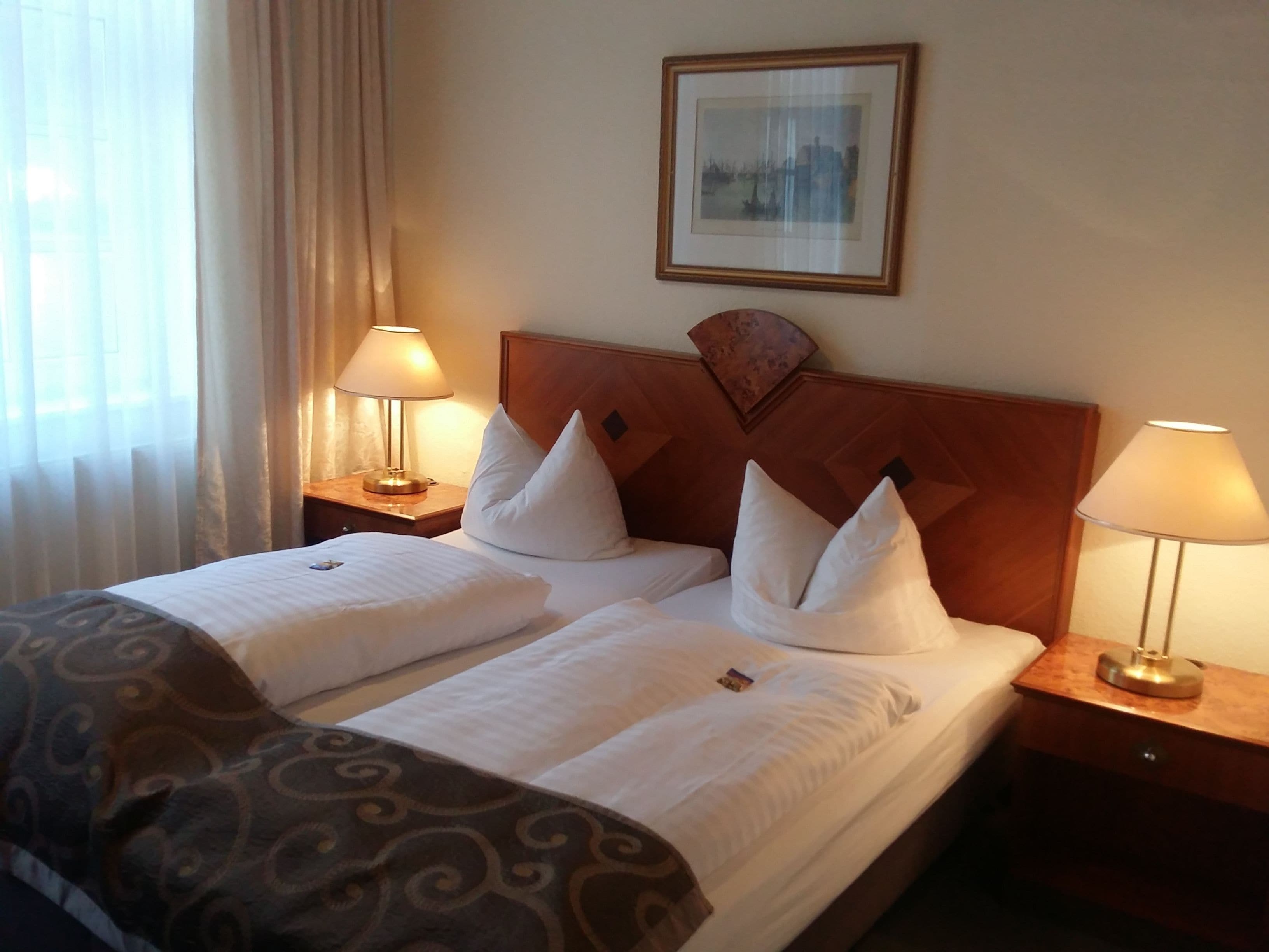 Habitación de hotel con una cama doble, dos mesitas de noche con lámparas, un cabecero, un cuadro enmarcado en la pared y una ventana con cortinas.