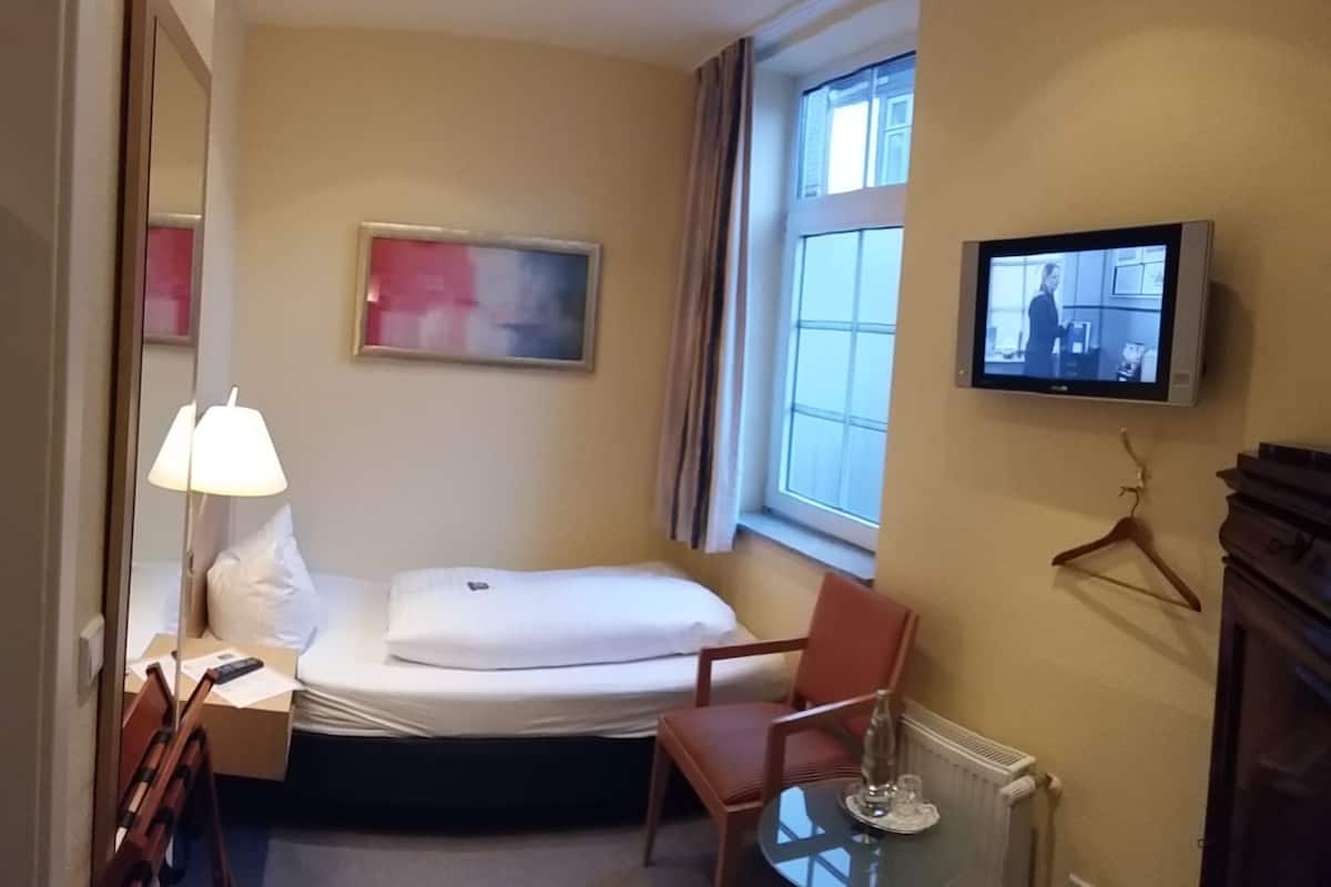 A hotel room with a bed, a chair, a table, a TV, a window, and a painting on the wall.