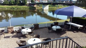 An outdoor patio with tables and chairs, a white picket fence, and a river in the background. - Goldener Anker (Bodenwerder)