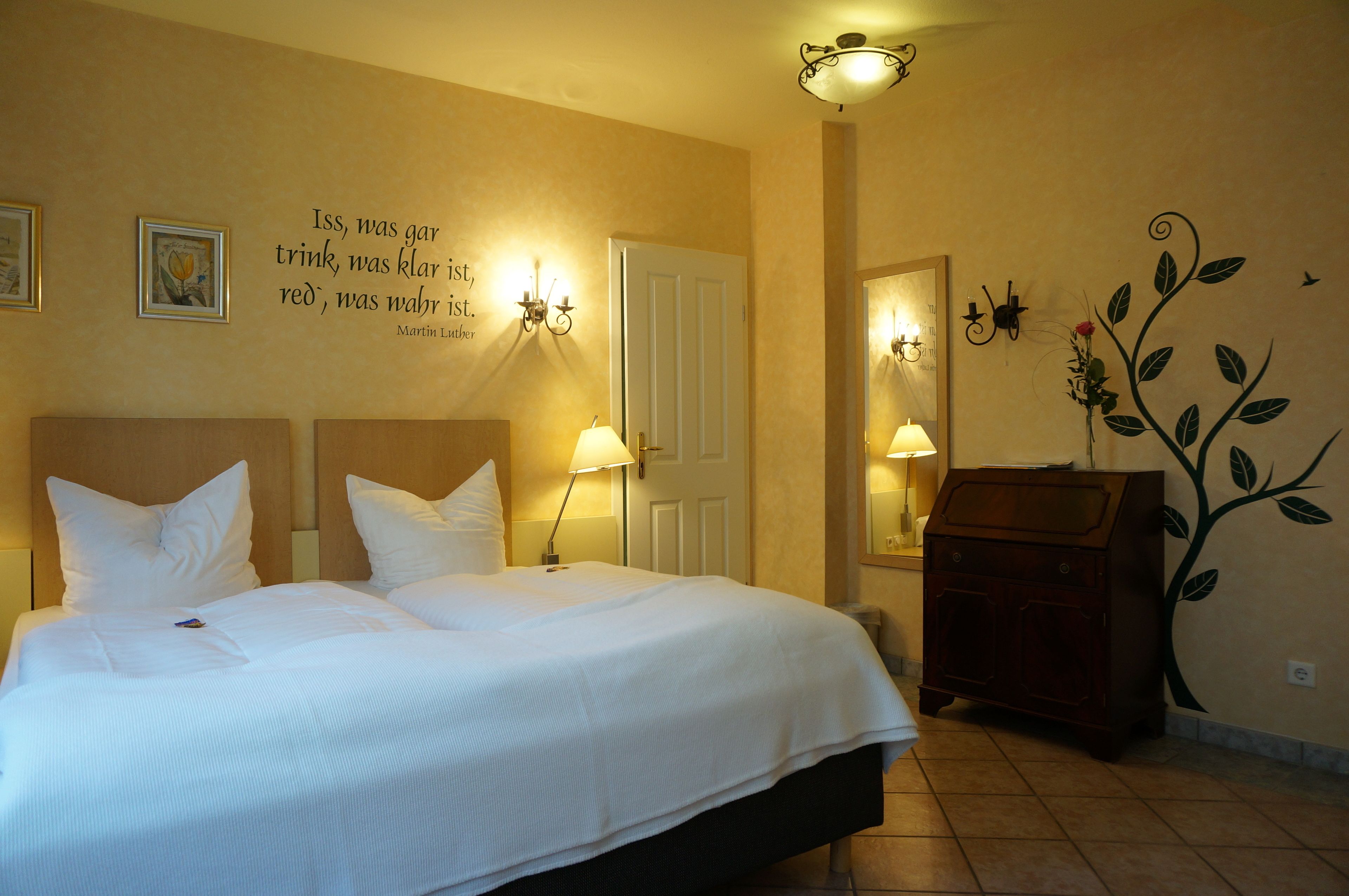 A hotel room with a large bed, two bedside lamps, a dresser, and a wall with a quote and a decorative plant.