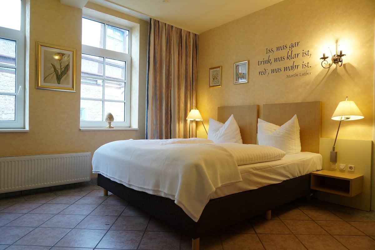 A hotel room with a large bed, two bedside lamps, a radiator, a window with curtains, and a quote on the wall.