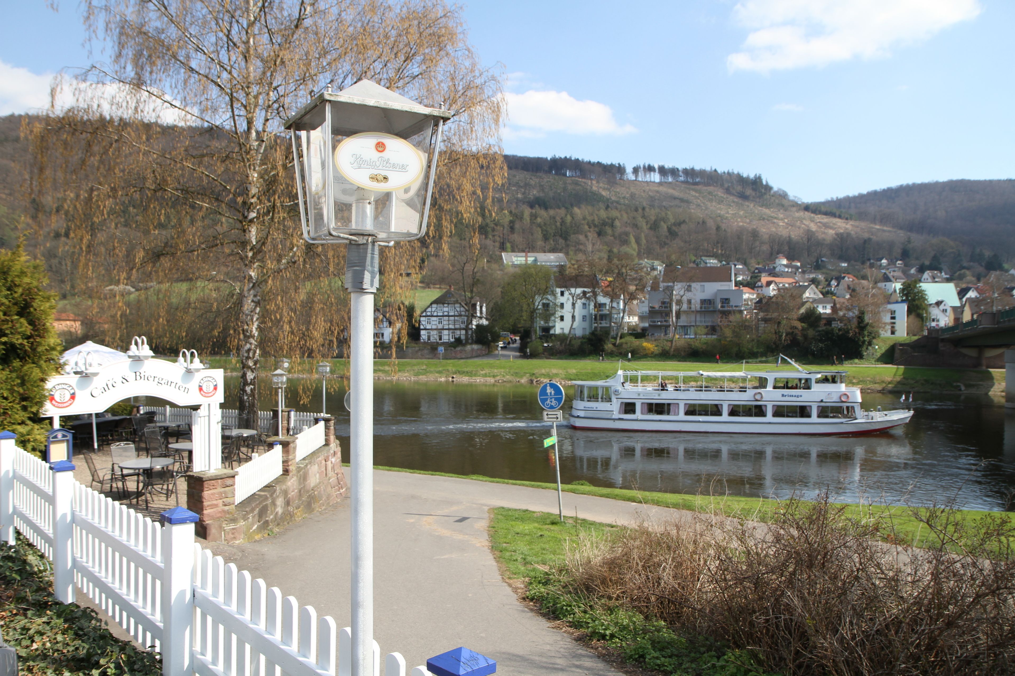 Uitzicht op een rivier met een witte boot, een café met terras en een witte hekken.