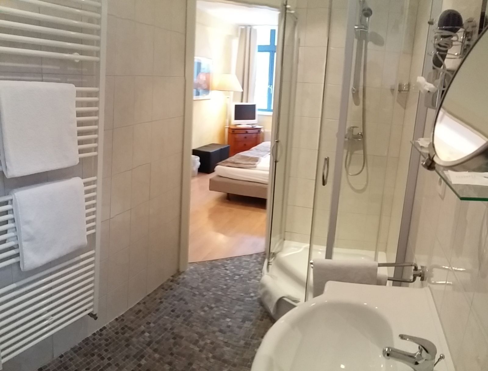 A bathroom with a shower, a sink, and a toilet. There is a bed visible in the background.