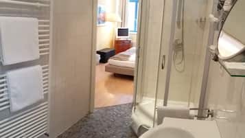 A bathroom with a shower, a sink, and a toilet. There is a bed visible in the background.