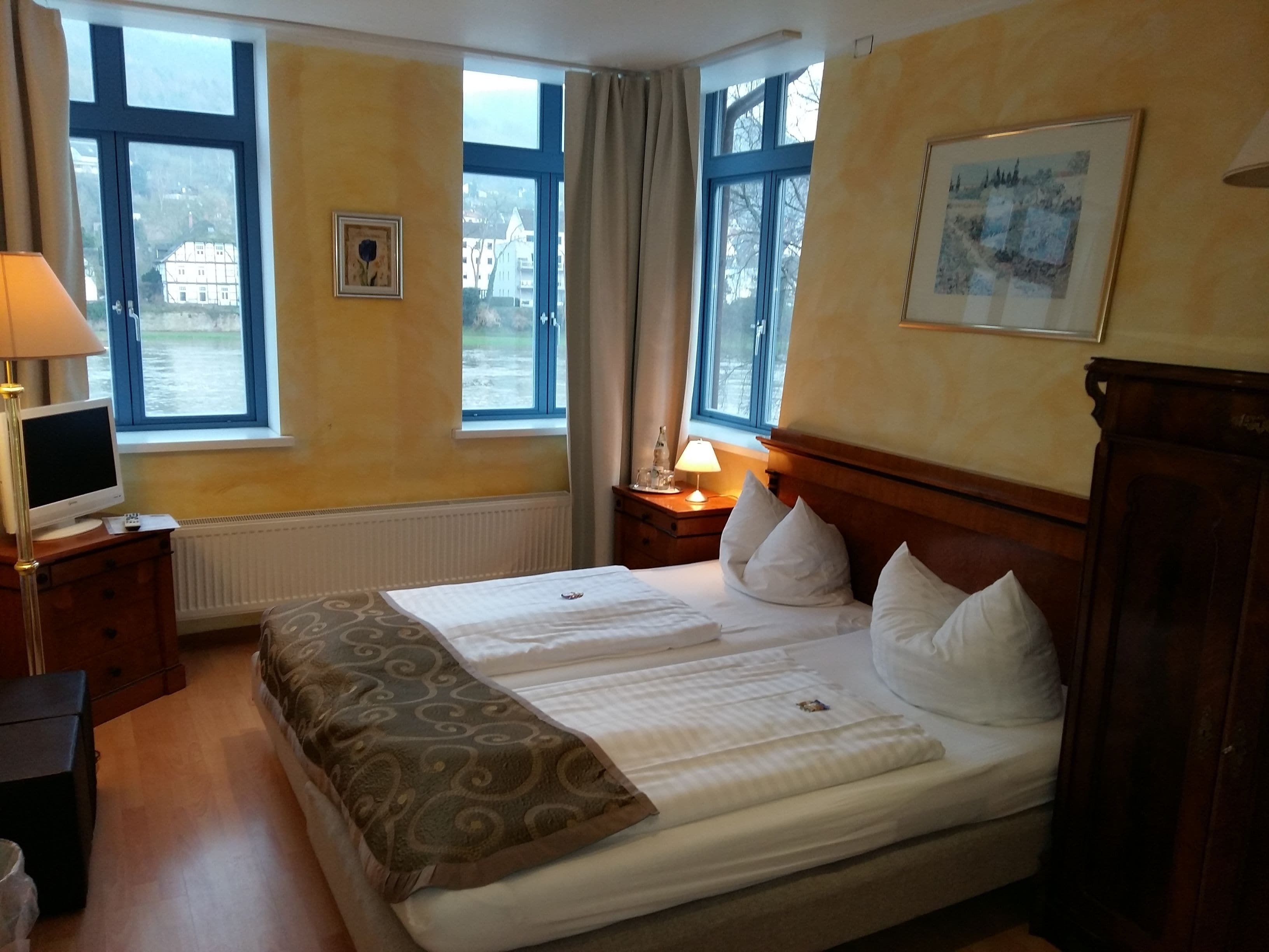 A hotel room with a bed, a TV, a lamp, and two windows.