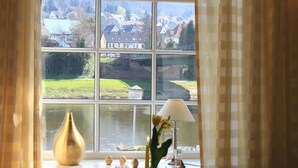 A dining table set with a vase of flowers, a lamp, and a view of a lake and houses through a window. - Goldener Anker (Bodenwerder)