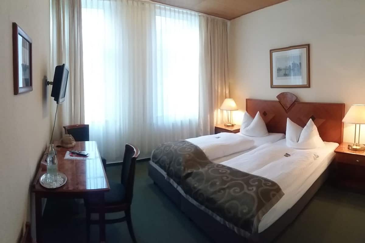 A hotel room with a bed, two bedside tables with lamps, a desk with a chair, a TV, and a window with curtains.