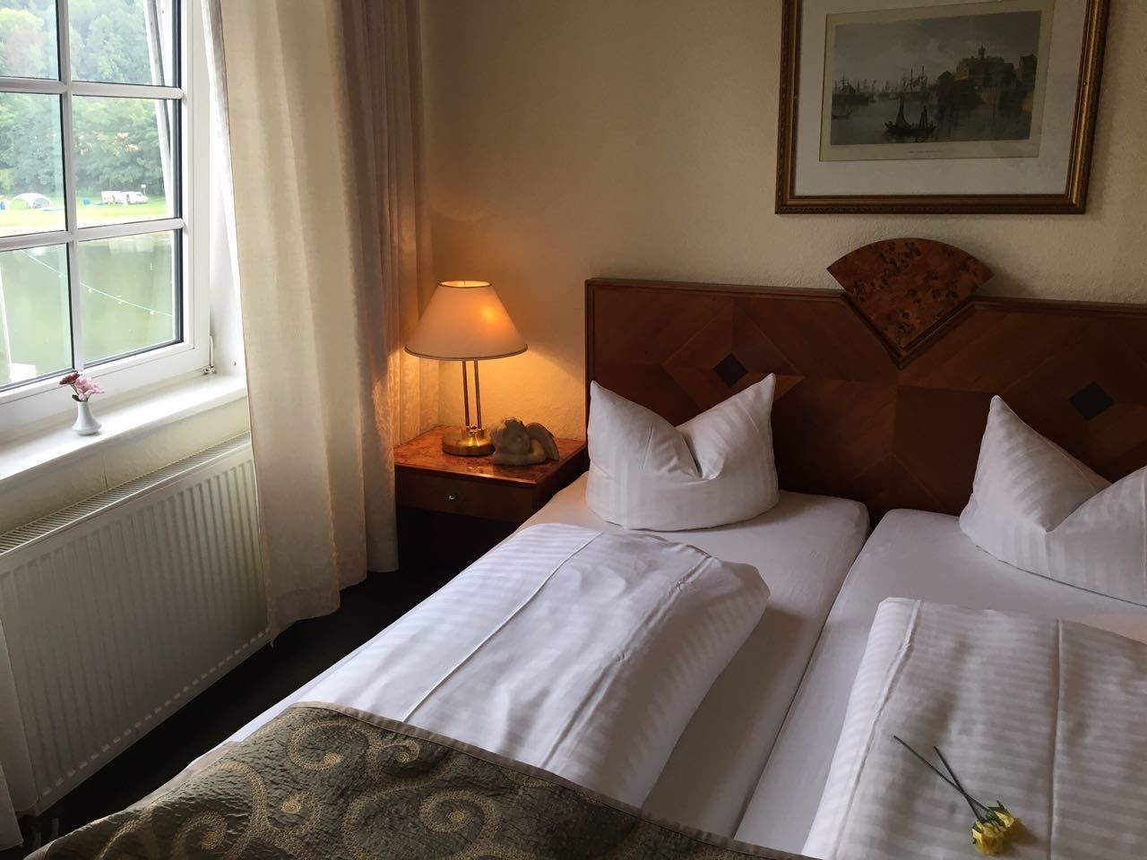 Una habitación de hotel con dos camas, una mesita de noche con lámpara, un cuadro en la pared y una ventana con cortinas.