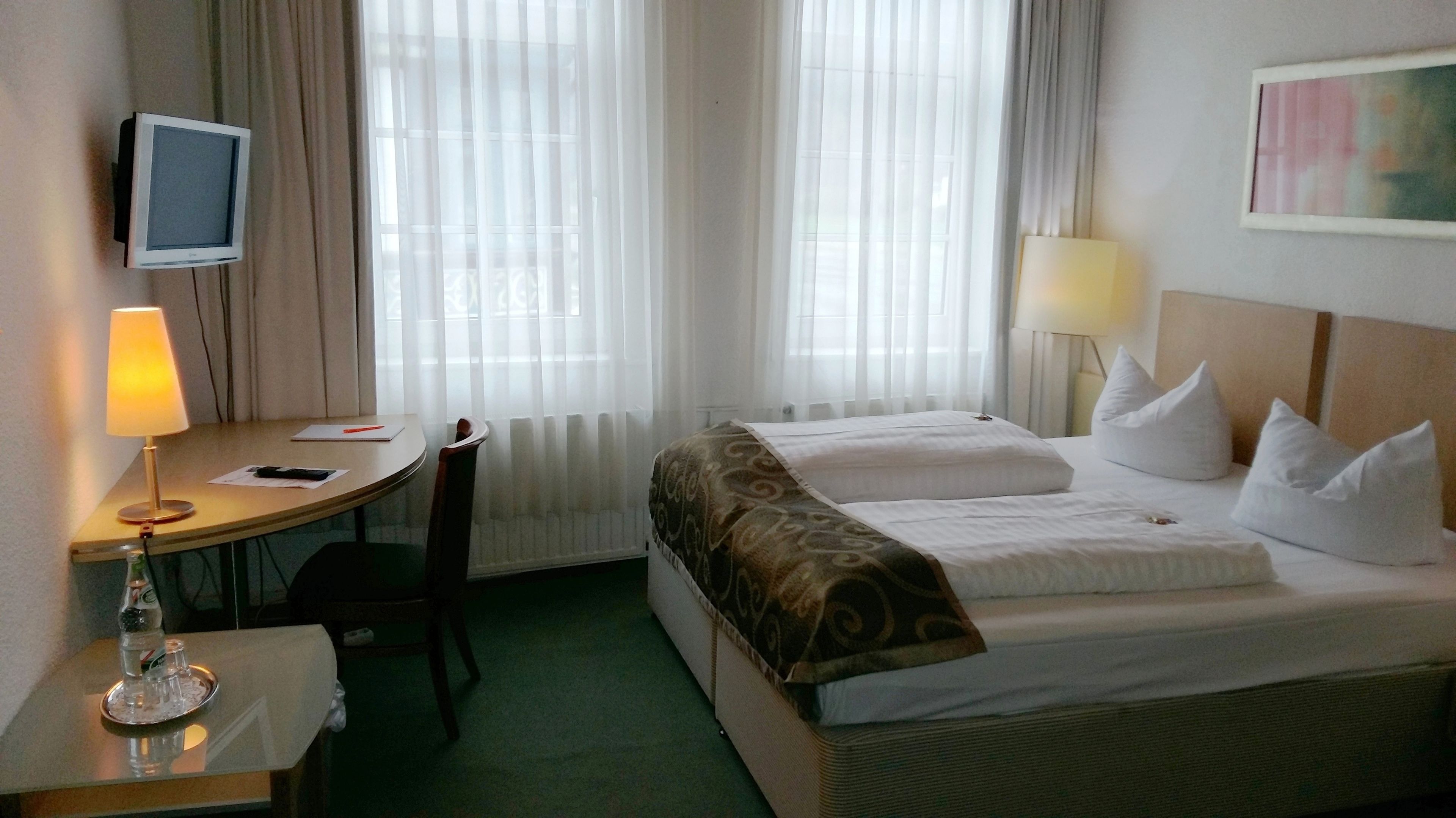 Une chambre d’hôtel avec un lit, un bureau avec une lampe, une télévision et une fenêtre avec des rideaux.