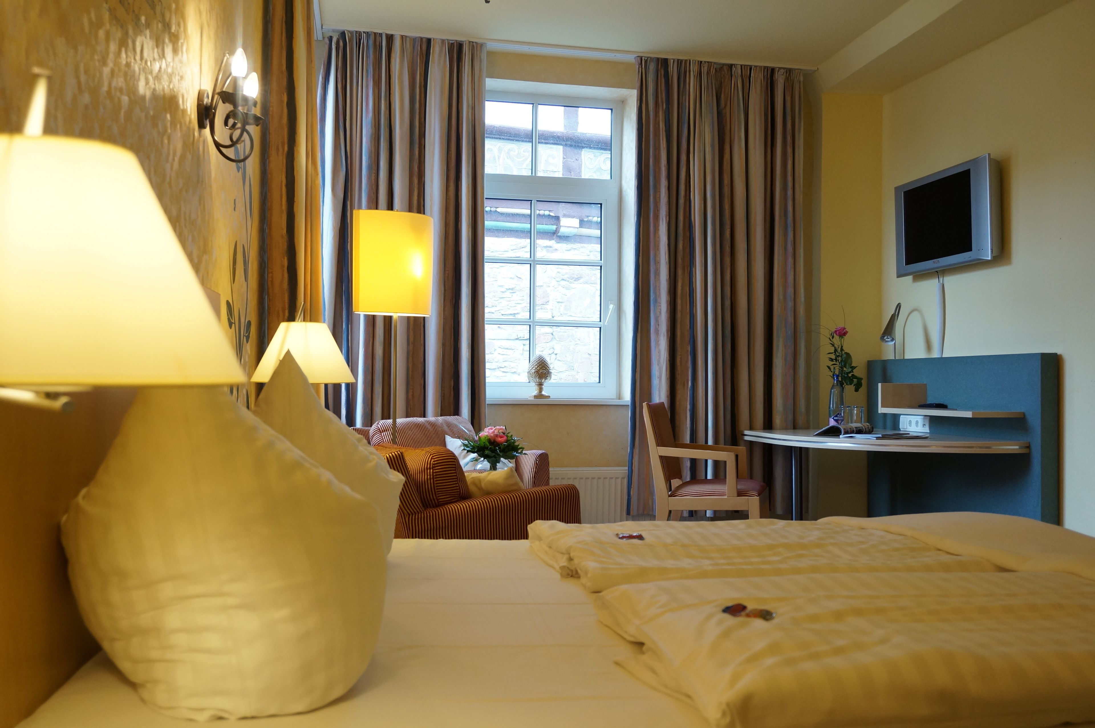 A hotel room with a bed, a desk, a chair, a lamp, a window with curtains, and a television.