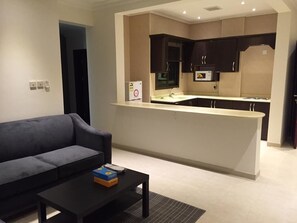 Un salon moderne avec un canapé et une table basse, une cuisine avec des meubles sombres et un plan de travail blanc, et un petit îlot central équipé d’un four à micro-ondes.