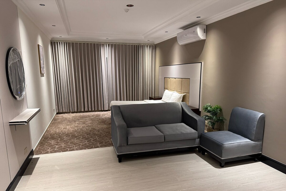 Une chambre d’hôtel moderne avec un canapé gris, un lit doté d’une tête de lit, un grand miroir et une plante en pot.