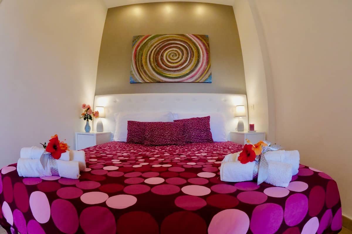 A bedroom with a circular patterned bedspread, a large abstract painting above the bed, and two bedside tables with lamps and flower arrangements.