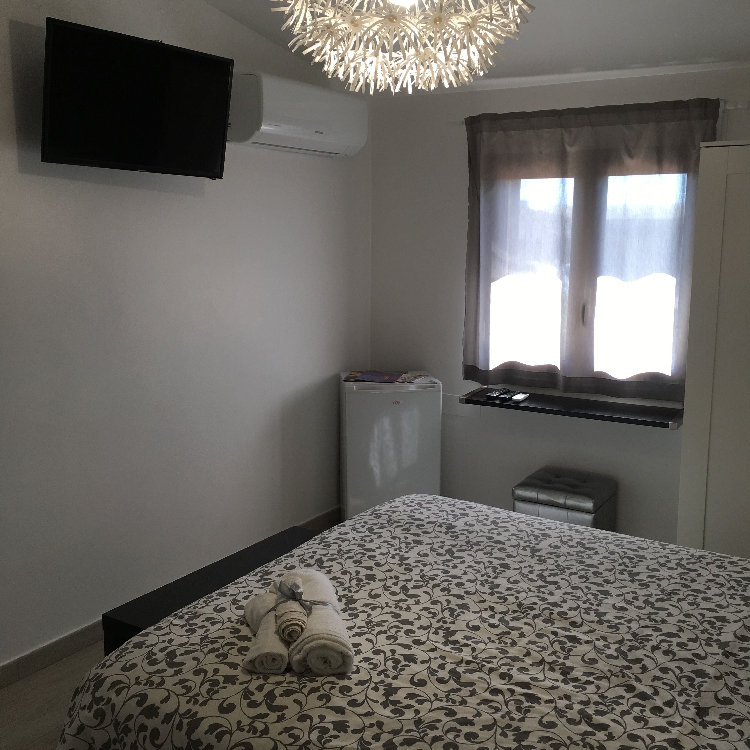 A bedroom with a bed, a TV mounted on the wall, an air conditioner, a window with curtains, and a small table with items on it.