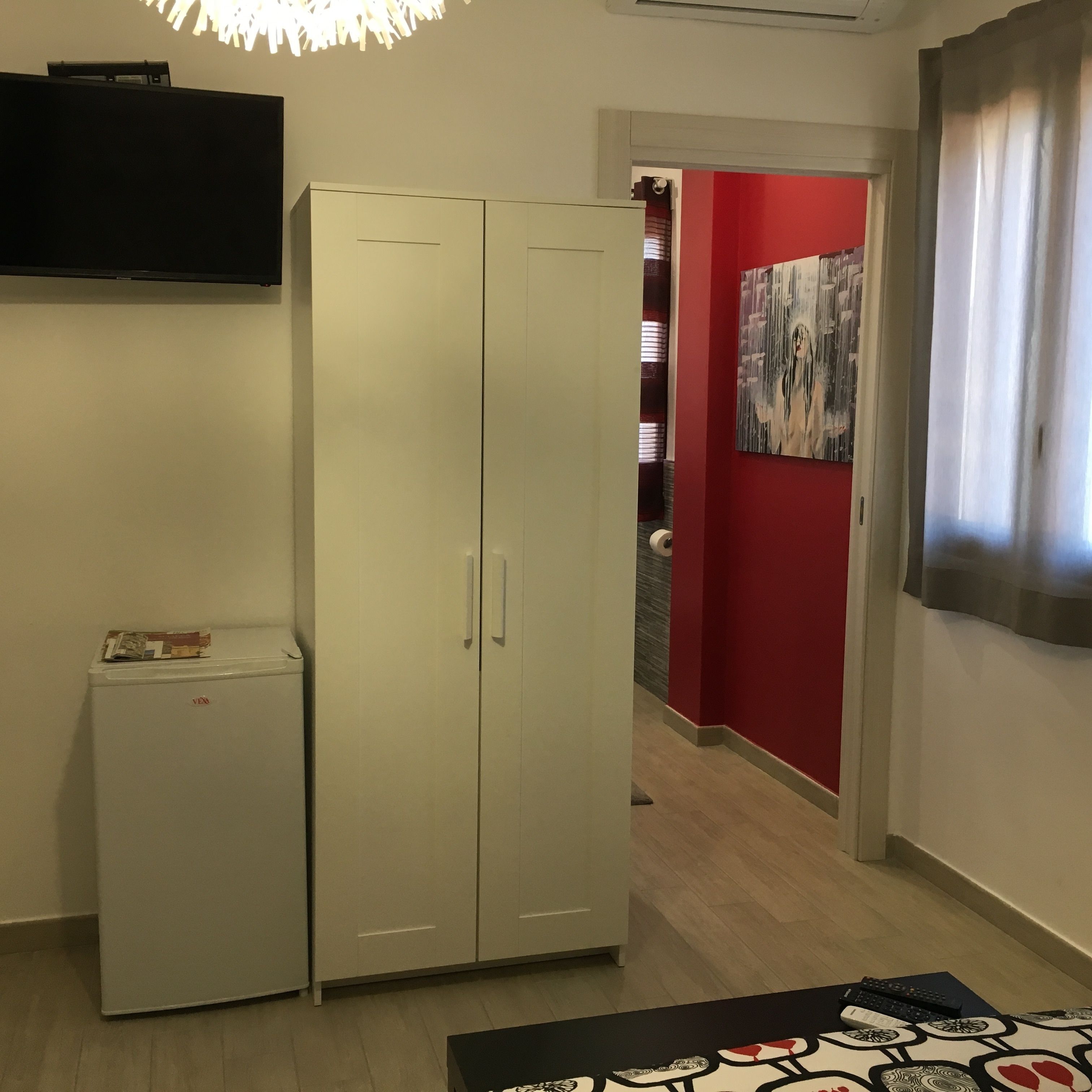 Una habitación con un armario blanco, un refrigerador pequeño, una pared roja y una puerta que da a otra habitación.