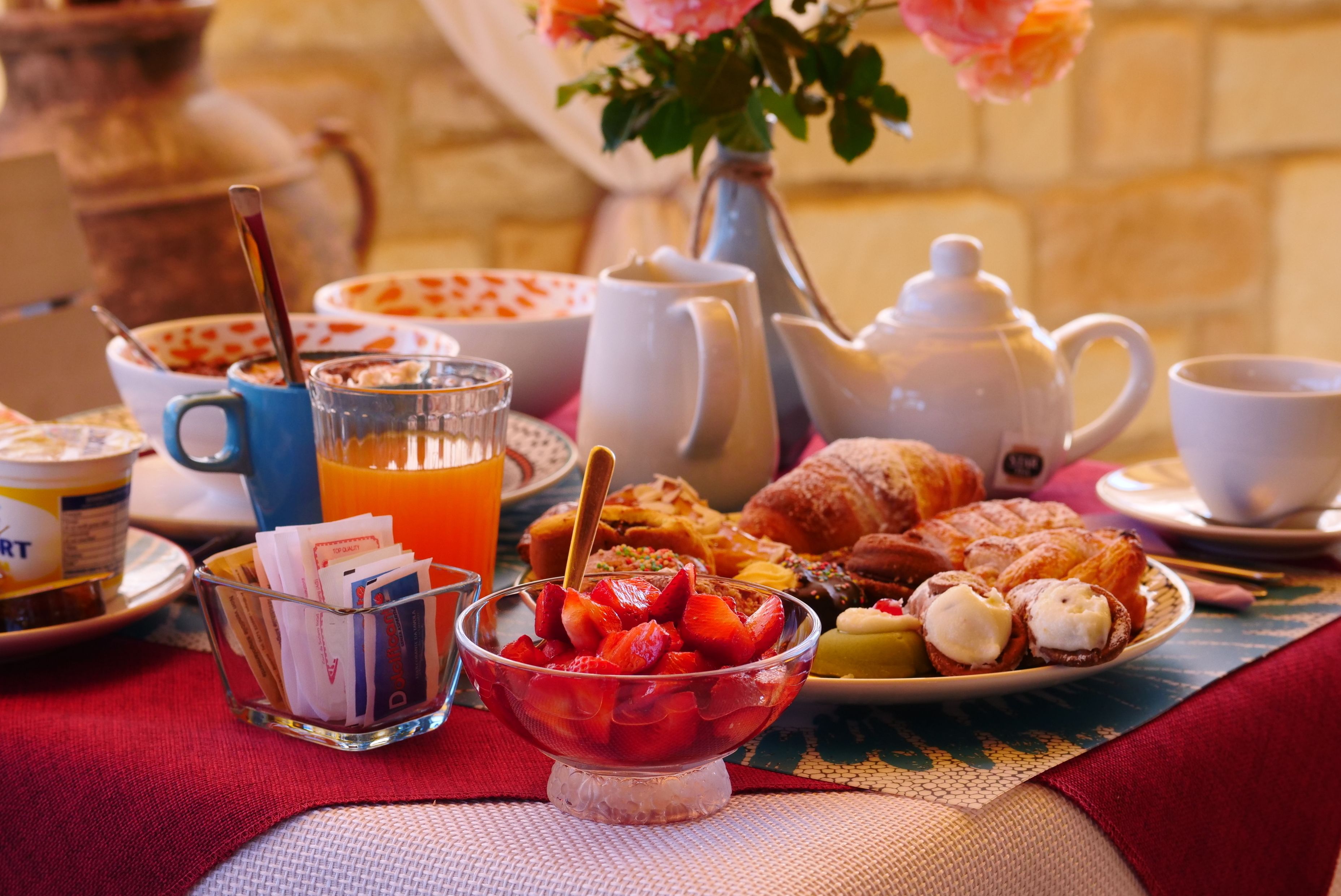A breakfast setting with a variety of food items, including pastries, fruit, and beverages, arranged on a table with a red tablecloth.