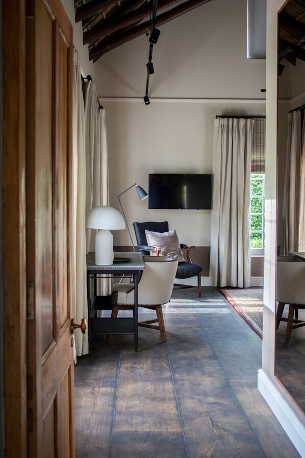 Quarto com piso de madeira, uma escrivaninha com luminária, uma cadeira, TV fixada na parede e vista para o verde lá fora.