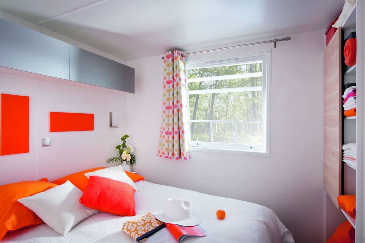 Een slaapkamer met een bed, kussens, een hoed, een boek en een raam met gordijnen.