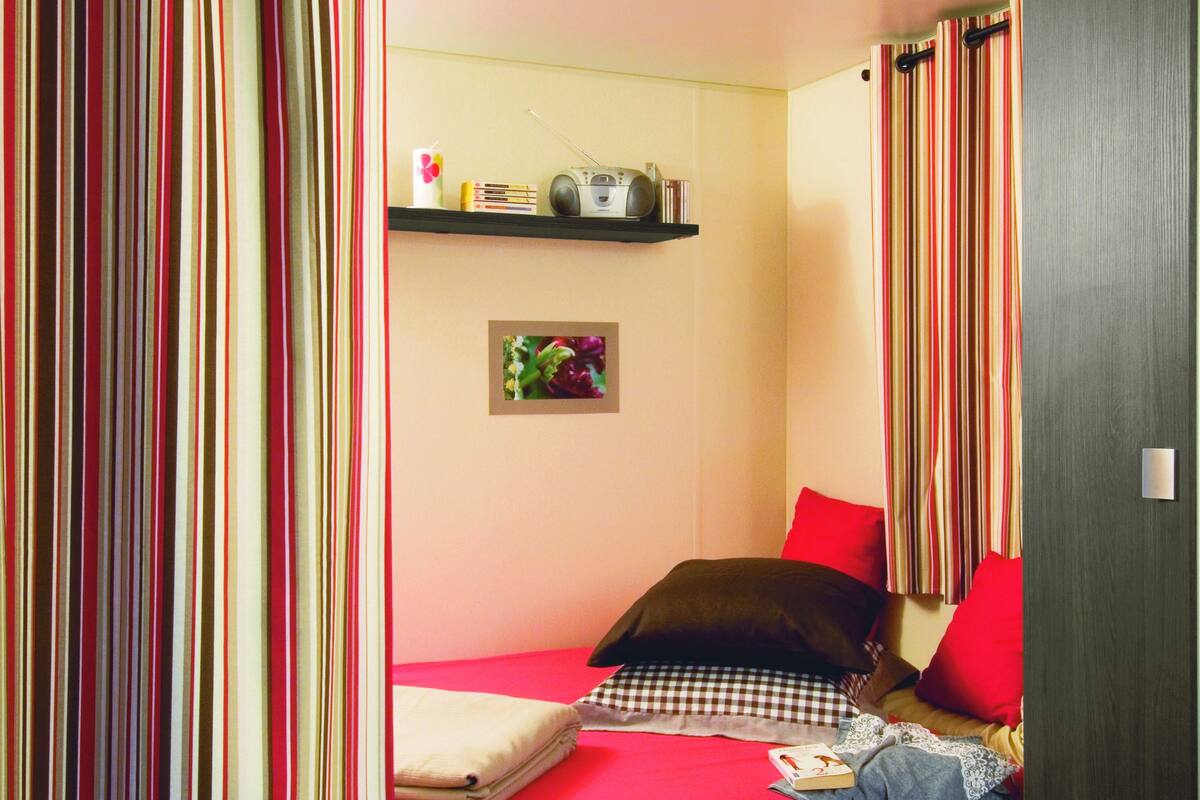 Een kleine, compacte kamer met een enkel bed, een plank met boeken en een radio, en een paar schoenen op de vloer.